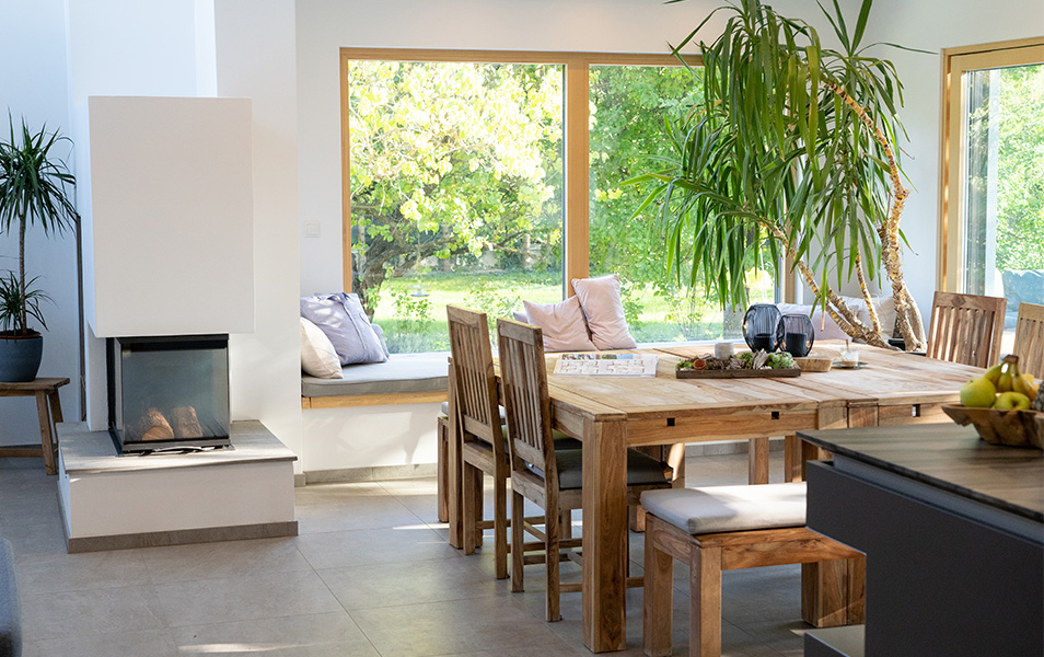 Modernes Wohnzimmer in einem schl&uuml;sselfertigen Fertighaus in &Ouml;sterreich mit gro&szlig;em Holztisch, Kamin und gro&szlig;en Fenstern, die viel Tageslicht hereinlassen.