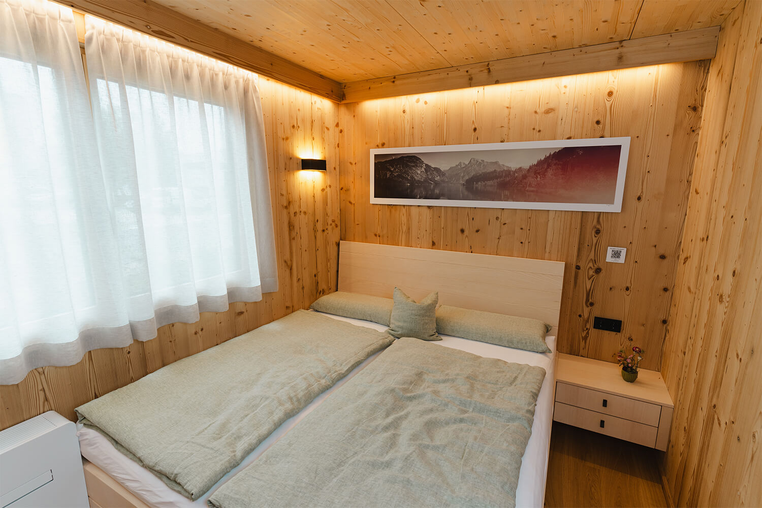 Schlafzimmer im individuell konfigurierten Tiny Haus von WOLF Haus mit gem&uuml;tlichem Doppelbett, heller Holzverkleidung, stimmungsvoller Beleuchtung und Bergpanorama-Wandbild